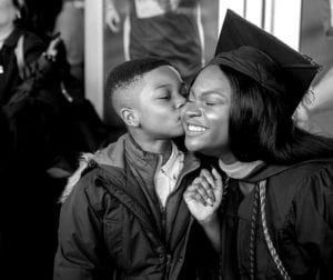 A woman in graduation gear being kissed by her young son